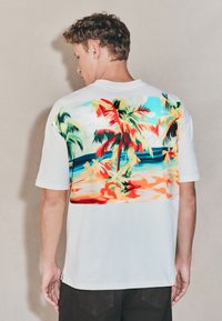 Vit t-shirt med korta ärmar med en färgglad strand- och palmträdsgrafik på baksidan, med en mjuk tygstruktur och avslappnad passform.