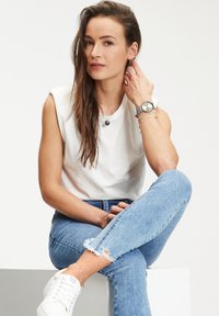 Witte mouwloze t-shirt, lichtblauwe distressed jeans, witte sneakers, zilveren horloge en gelaagde armbanden. Eenvoudige, casual outfit.