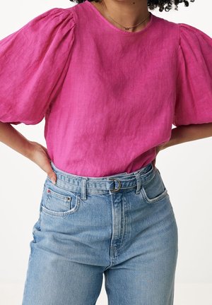 Kvinde iført en lyserød bluse med pufærmer, stoppet ned i højtaljede lyseblå jeans med bælte, hænderne hvilende på hofterne.
