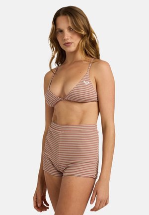 Femme aux cheveux ondulés portant un haut de bikini rayé et un short taille haute dans des tons beige et rouge sur un fond uni.