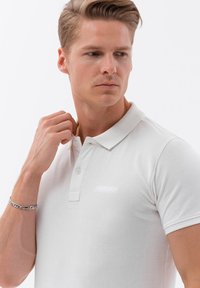 Bílá polo košile s texturovanou látkou, která má dvojité zapínání na knoflíky a skládací límec. Model upravuje límec, ukazující volný střih.