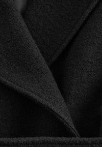 Schwarzer Wollmantel mit strukturierter Oberfläche, ausgestattet mit einem breiten Revers und überlappendem Stoff, der Tiefe und Struktur betont. Keine sichtbaren Verschlüsse.