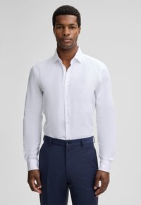Man staat rechtop met het gezicht naar voren, gekleed in een witte lange mouwen overhemd en marineblauwe pantalon tegen een eenvoudige grijze achtergrond.
