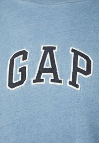 Himmelblaues Sweatshirt aus Baumwolle mit einer strukturierten Textur, das auf der Vorderseite das Wort "GAP" in großen, dunkelblauen, gestickten Buchstaben zeigt.