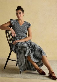 Junge Frau mit einem hohen Dutt sitzt auf einem Holzstuhl, trägt ein schwarz-weißes Karokleid und burgunderrote Schuhe mit Doppelriemen vor einer beigen Wand.