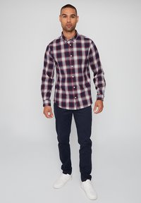 Camisa de cuadros en rojo, azul marino y gris con cuello abotonado, combinada con pantalones azul marino oscuros y zapatillas blancas. Textura suave de algodón.