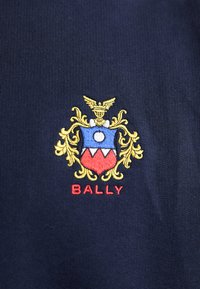Il tessuto blu navy presenta un logo ricamato con uno scudo blu, dettagli dorati, un cerchio bianco, elementi rossi e il testo "BALLY" in rosso.