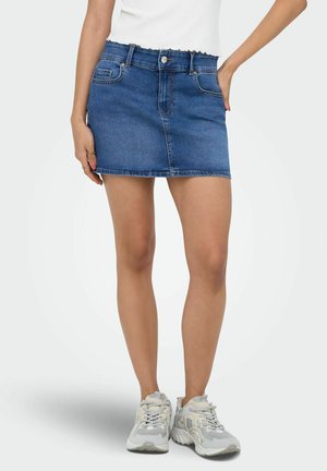 Kobieta ubrana w niebieską mini spódniczkę z denimu z kieszeniami, biały top i białe siatkowe tenisówki, stojąca z jedną ręką na biodrze.