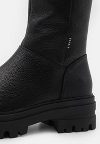 Botín de cuero negro texturizado con suela de goma gruesa y un pequeño logotipo en el lado. Presenta una forma suave y redondeada y una construcción duradera.