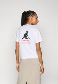 Valge lühikeste varrukatega T-särk, millel on must kenguru graafika ja "KANGOL J.LINDEBERG" tekst roosa ja oranži värviga seljal.