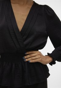 Svart blus med V-ringning, plisserat tyg och smockad midja. Långa, puffiga ärmar med volangslut. Slätt, något texturerat material.