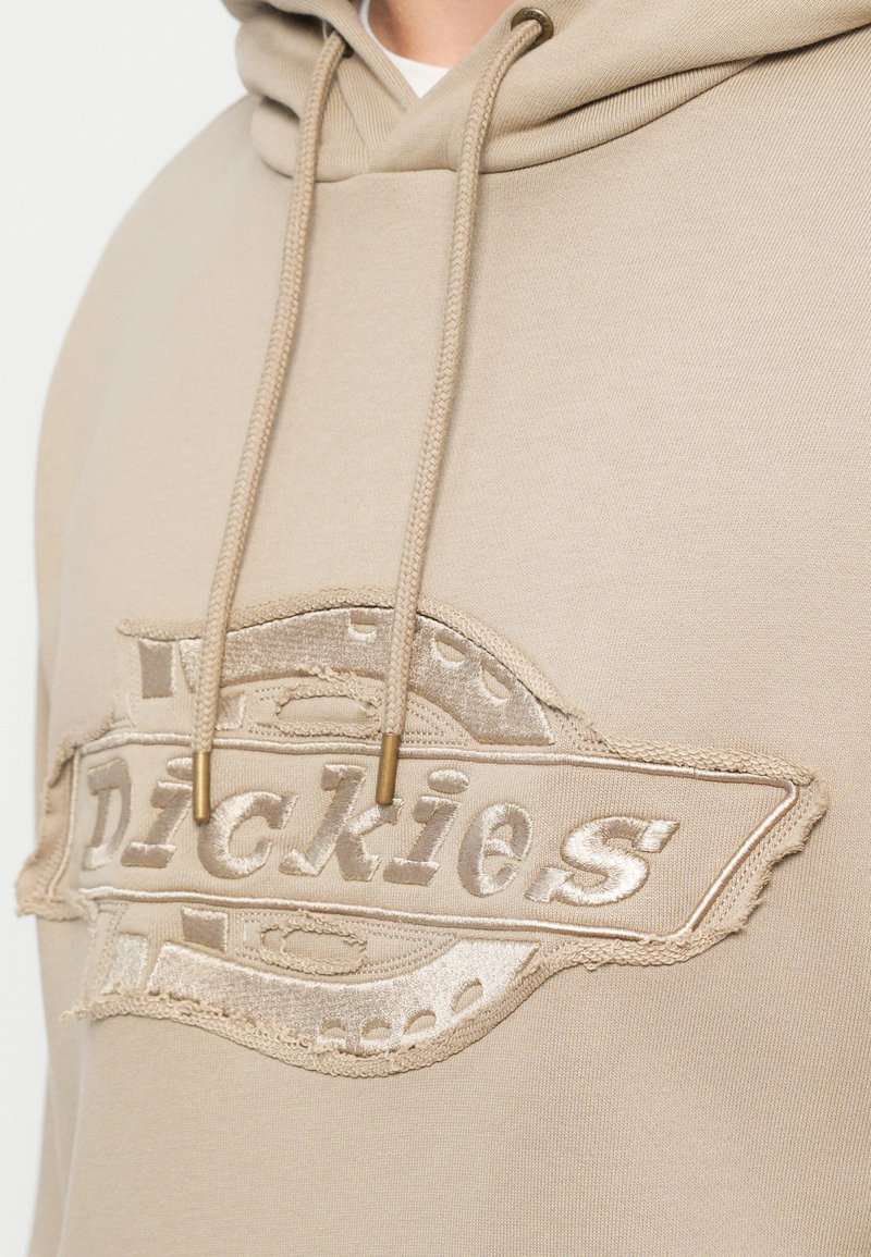 Beige hoodie with a textured, distressed logo patch. Hood features matching drawstrings with metal tips. Soft, cotton blend fabric.