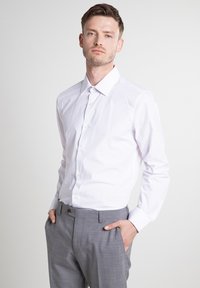 Bela srajca z dolgimi rokavi s klasičnim ovratnikom, ki ima gladko teksturo in zapiranje na gumb, v kombinaciji s svetlo sivimi vzorčastimi hlačami.