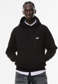 Felpa nera con cappuccio realizzata in tessuto, con una tasca frontale a marsupio e un piccolo logo bianco sul petto. Indossata sopra una camicia con colletto.