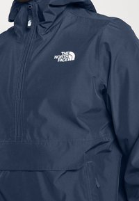 Giacca leggera, impermeabile, di colore blu navy, con zip a metà e cappuccio regolabile, e un logo bianco prominente sul petto.