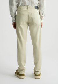 Pantalones casuales color beige claro con un diseño de pierna recta, bolsillos traseros y un parche de cuero con el logotipo en la cinturilla. Llevados con una camisa a rayas.