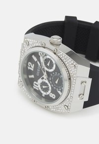 Reloj en tono plateado con esfera negra, grandes números, funciones de cronógrafo y adornos de cristal. Correa de goma con detalle texturizado.