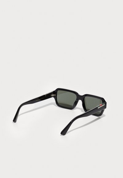 Lunettes de soleil noires rectangulaires avec verres verts, monture en plastique épaisse et accents subtils sur les branches. Présente un logo sur le côté gauche.