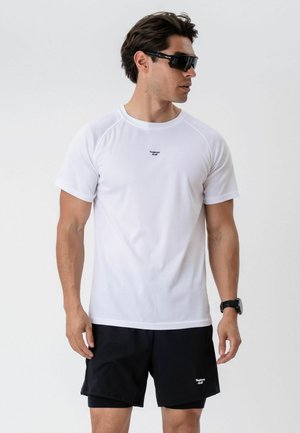 Man met een wit T-shirt met korte mouwen en zwarte korte broek, zwarte horloge en zwarte sportzonnebril, staand tegen een effen witte achtergrond.