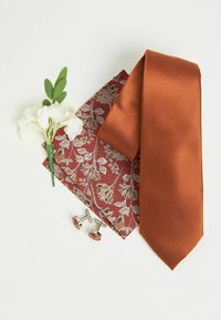Cravate satin orange, pochette de costume florale maroon, boutons de manchette en argent avec un motif floral, et une petite fleur blanche avec des feuilles vertes.