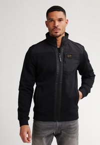 Chaqueta negra con cremallera y cuello alto, de tela texturizada, con bolsillo en el pecho y detalles de tela contrastante en los hombros. Combinada con jeans grises.