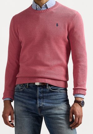 Mies, joka pitää yllään vaaleanpunaista neulepuseroa sinisen napitettavan paidan päällä, siniset farkut ja ruskean nahkahihnan rannekellon vasemmassa ranteessa.