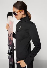Giacca da sci nera con colletto alto e branding discreto, abbinata a sci rosa con accenti neri e grigi. Gli accessori includono occhiali da sole.