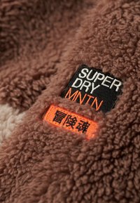 Brązowy, puszysty materiał polarowy z czarnymi i pomarańczowymi haftowanymi naszywkami z napisami "SUPER DRY" oraz "MNTN" z boku. Tekstura wydaje się miękka i gruba.
