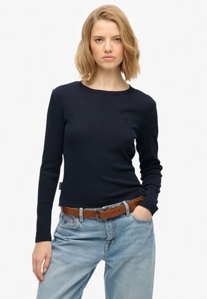 Mujer joven que lleva una camiseta ajustada de manga larga azul marino, vaqueros azul claro y un cinturón de cuero marrón, de pie con una mano en el bolsillo.