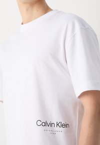 Biała bawełniana koszulka z okrągłym dekoltem, krótkimi rękawami i czarnym nadrukiem logo z napisem "Calvin Klein" oraz "ESTABLISHED 1968" na dolnym brzegu.