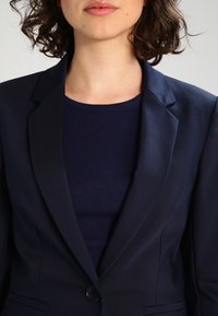 Námořnický blazer s přiléhavým střihem, výstřihem do "V", zapínáním na jeden knoflík a hladkou texturou, vrstvený přes námořnický top s kulatým výstřihem.
