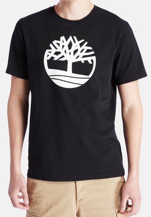 Mann trägt schwarzes Kurzarm-T-Shirt mit großem weißem stilisiertem Baumlogo auf der Brust und beiger Hose vor weißem Hintergrund.