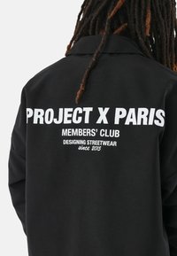 Chaqueta negra con una amplia impresión blanca en la parte posterior que dice "PROJECT X PARIS MEMBERS' CLUB" y texto adicional sobre diseño streetwear.