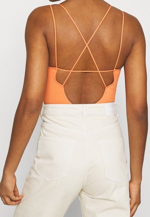 Person iført hvide bukser med høj talje og en orange ribbet tanktop med tynde, krydsede stropper og en åben ryg.