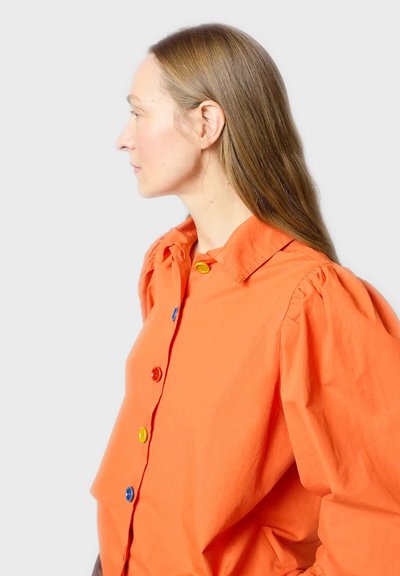 Oranssi puuvillapaita, jossa on puffihihat ja etupaneelissa moniväriset napit punaisena, keltaisena ja sinisenä. Sivukuva.