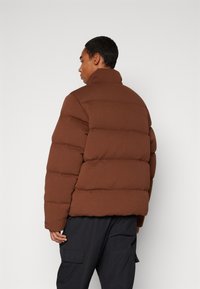 Giubbotto imbottito marrone con colletto alto, caratterizzato da un design trapuntato e una vestibilità leggermente ampia, indossato con pantaloni scuri.