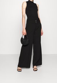 Černý top bez rukávů s texturovaným žebrováním, spárovaný s širokým černým kalhotami s vysokým pasem a dvojitými knoflíkovými detaily. Drží malou prošívanou černou tašku.