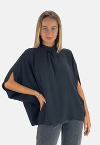 Schwarzes, leichtes Top mit hohem Kragen und gesplitteten Kurzärmeln, aus strukturiertem Stoff und mit entspannter Passform. Kombiniert mit grauen Jeans.