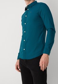 Turquoise lange mouwen shirt van gladde stof, met een button-down kraag, witte knopen en een subtiel logo op de borst. Gecombineerd met zwarte broek.