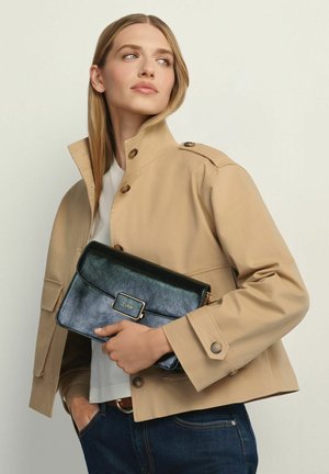 Kobieta o długich blond włosach, ubrana w beżową kurtkę i białą koszulę, trzymająca czarną skórzaną kopertówkę, patrząca w bok.