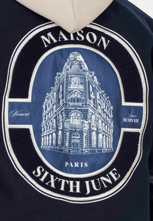 Marineblauwe hoodie met een gestructureerd rond patch waarin een witte gebouwillustratie is afgebeeld, met de woorden "MAISON", "PARIS" en "SIXTH JUNE" in contrasterende kleuren.