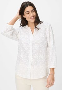 Weiße, knöpfbare Bluse mit floralem Stickerei, langen Ärmeln mit elastischen Bündchen, einem subtilen Glanz und einem abgerundeten Saum. Leichtes Material.