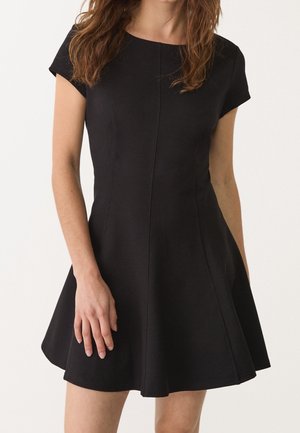 Vrouw draagt een nauwsluitende zwarte jurk met korte mouwen en een uitlopende rok, staand tegen een effen lichte achtergrond.