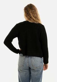 Zwarte gebreide trui met een ontspannen pasvorm, voorzien van lange mouwen en een cropped achterkant, gecombineerd met lichtblauwe, high-waisted jeans.