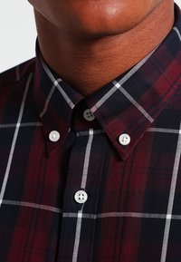Primo piano di una persona che indossa una camicia a quadri rossi scuri e blu navy con bottoni bianchi e colletto abbottonato.