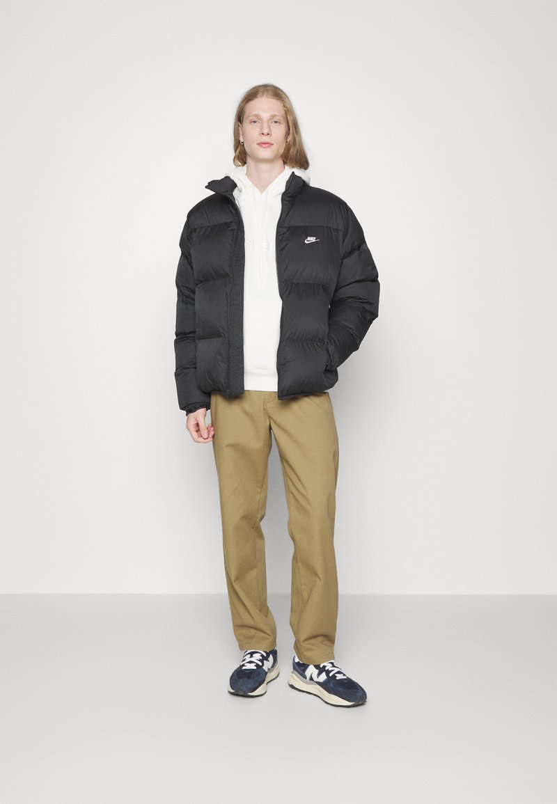 Chaqueta de plumas negra con costuras horizontales, logo blanco en el pecho, llevada sobre una sudadera blanca, combinada con pantalones caqui y zapatillas azul marino.