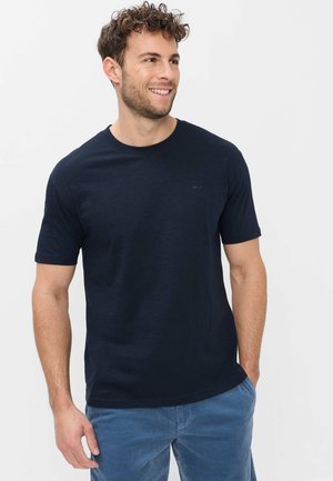 Mand med krøllet hår, der smiler, iført en sort kortærmet t-shirt og blå bukser i fløjl, stående med den ene hånd i lommen.