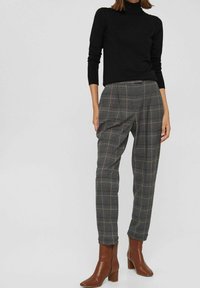 Zwarte coltrui met nauwsluitende mouwen. Grijze geruite broek met plooien en een losse pasvorm. Bruine enkellaarsjes met blokhakken.