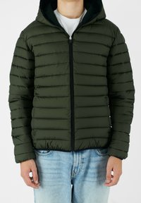 Grøn polstret jakke med hætte, vandret syninger, lynlås lukning og tætsiddende design. Båret over en hvid skjorte og lyseblå jeans.