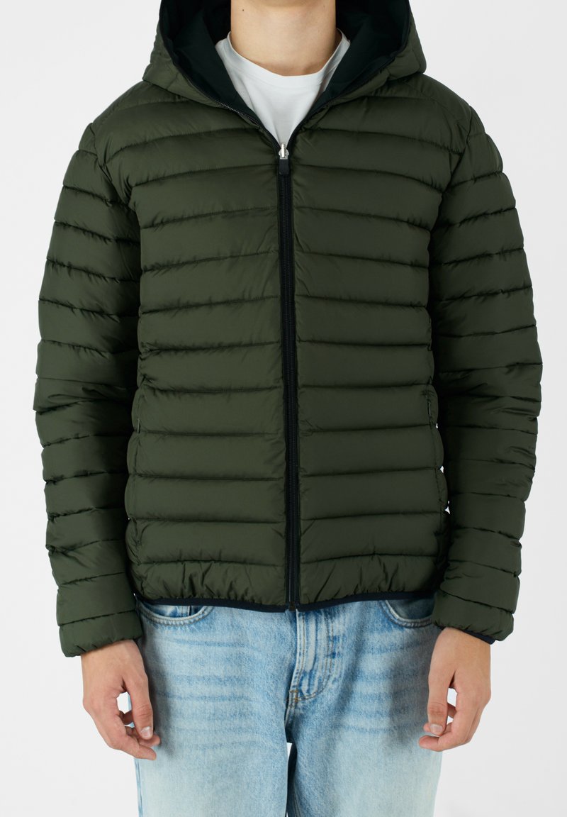Grøn polstret jakke med hætte, vandret syninger, lynlås lukning og tætsiddende design. Båret over en hvid skjorte og lyseblå jeans.